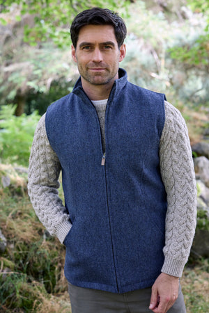 A man wearing a navy blue zip-up fleece vest over a light gray cable-knit sweater, standing outdoors in a natural, grassy environment.